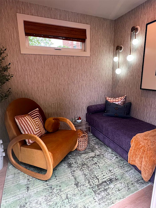 A photo of a cozy interior of the SheCare Wellness Pods Resting Room. With a carpeted floor and soft colored walls, this Resting Pod features a lounge bed with pillows and blanket, as well as a velvet lounge chair. Modern art adorns the walls, accented by soft warm lighting.