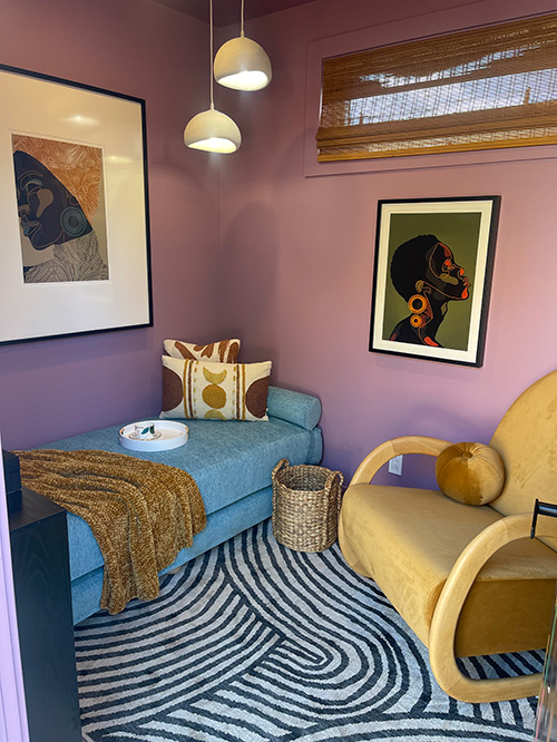 A photo of a cozy interior of the SheCare Wellness Pods Resting Room. With a carpeted floor and soft colored walls, this Resting Pod features a lounge bed with pillows and blanket, as well as a velvet lounge chair. Modern art adorns the walls, accented by soft warm lighting.