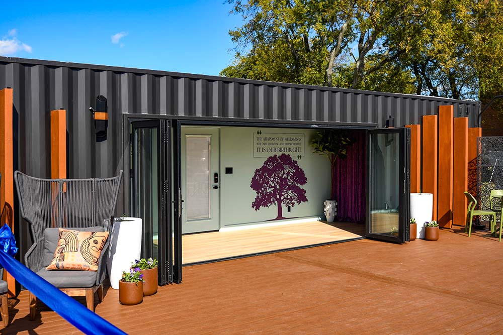 A photo of one of the two SheCare Wellness Pods exterior. A repurposed freight container that has been painted and renovated into a outdoor building. Wooden art sculptures stand on either side of the opened, large, glass doors. In front of the doors is a large, open deck patio.