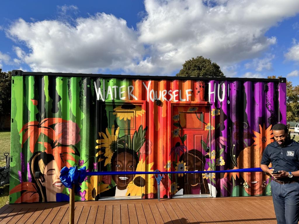 A photo showing the front view of one of the SheCare Wellness pod. A repurposed freight container painted with vibrant colors and depictions of young women smiling surrounded by flowers. The words "Water Yourself, HU!" are displayed about the two installed doors.