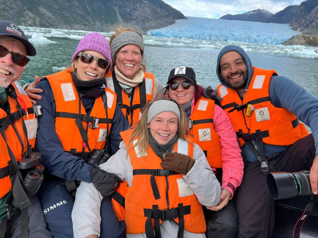 A photo of a professor and several students on a boat wearing orange lifevests with a large iceberg in the background.