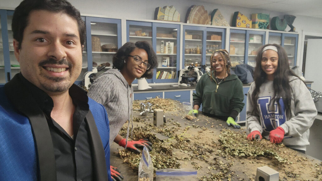 A photo of a professor and three students examining pieces of plants on a table.