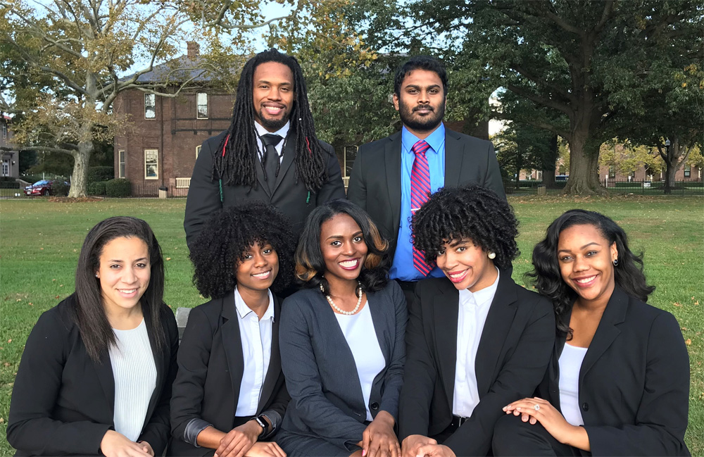 A photo of seven students on the OGS Executive Team wearing business professional attire while posing together for the camera outdoors on Hampton University's campus.