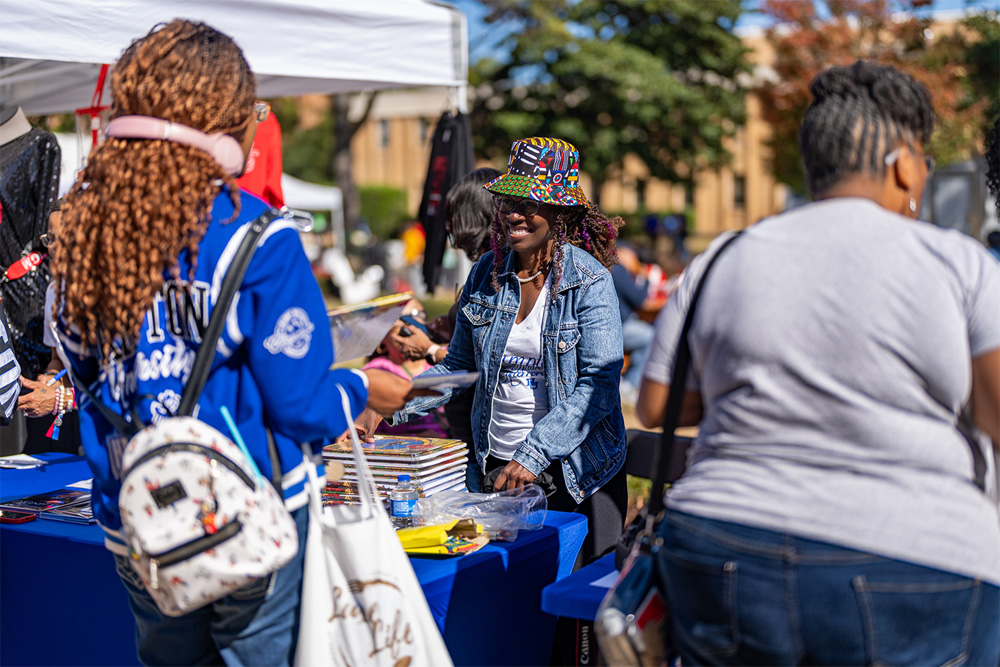 A photo of a woman wearing a Hampton University jacket buying something from a vendor's stall.