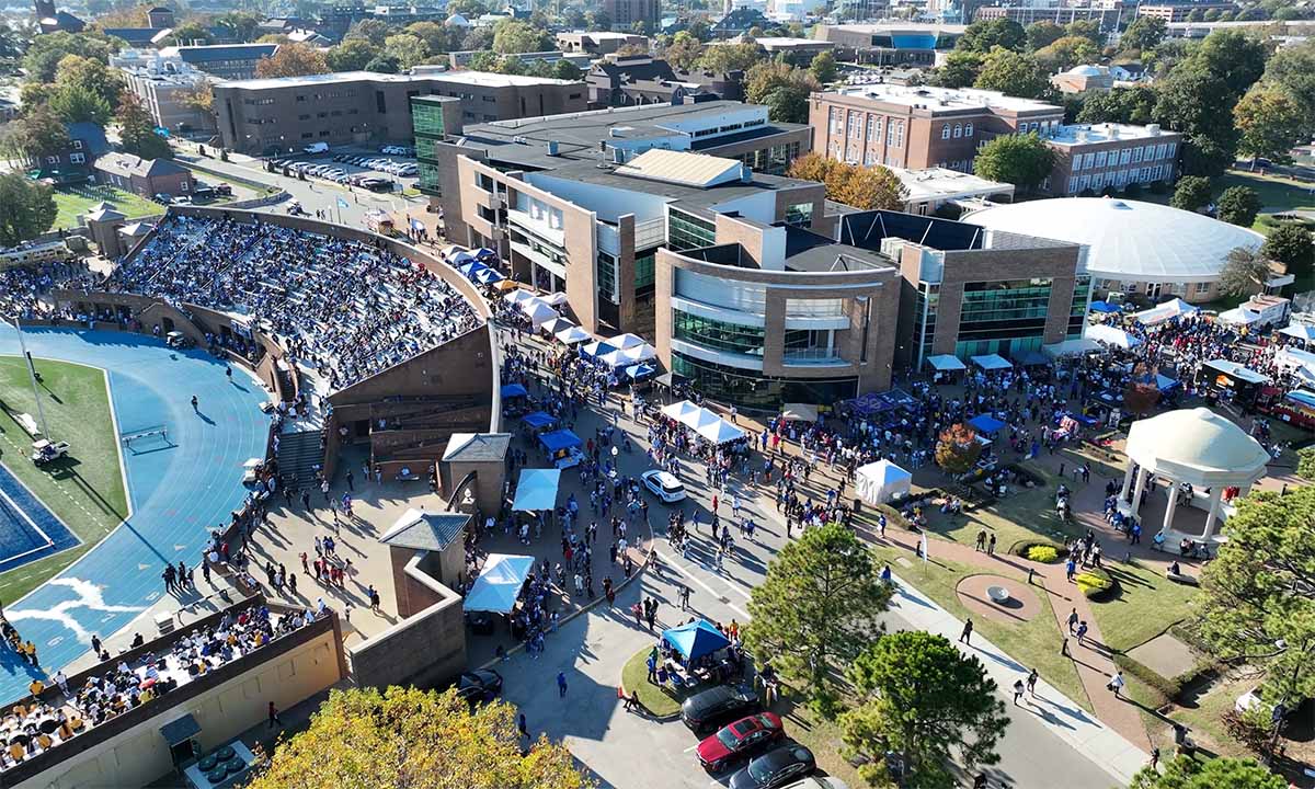 A photo taken from an aerial drone showing the Hampton University campus bustling with activity from a large crowd. The photo includes the Armstrong Stadium and the Student Center.