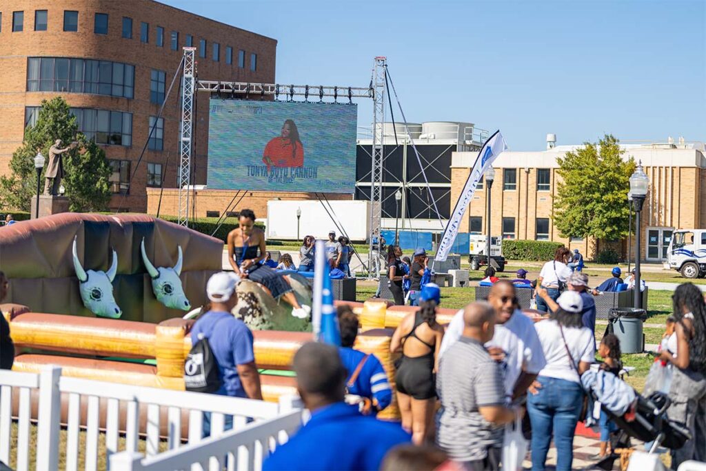 A photo from the Homecoming Tailgate. The photo was taken on the Booker T. Washington Memorial lawn behind the School of Pharmacy. There is a large screen showing music videos while a young woman rides a mechanical bull.