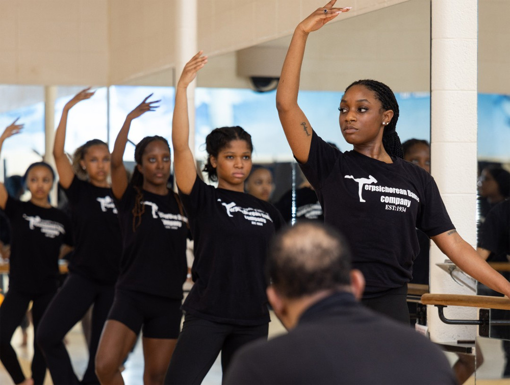 A photo of dance students posing in front of a mirror with one of their hands on a support beam.