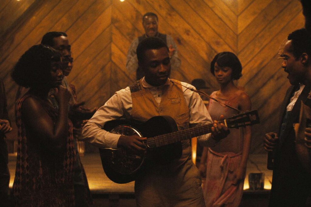 A screenshot of the movie "Sinners". People are standing in a darkly lit area while a man in the middle plays a guitar.