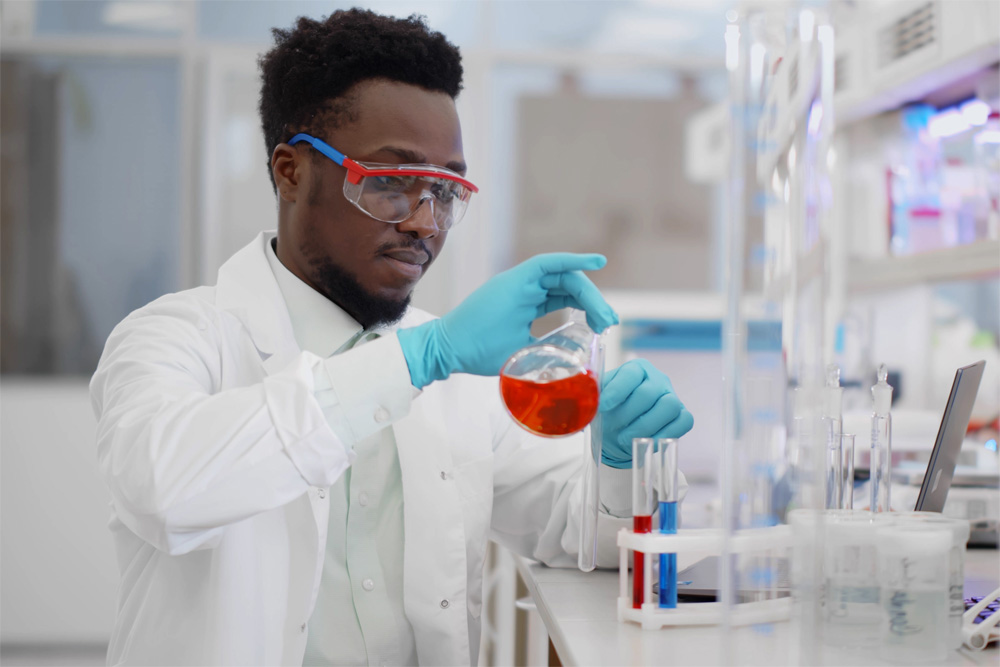 Photo of a man in a lab coat pouring a beaker with red fluid into an empty test tube.