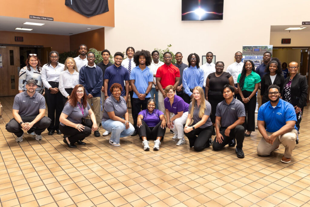 A large group of students poses shoulder to shoulder for the camera as a group of Leidos representatives crouch pose in front of the students.