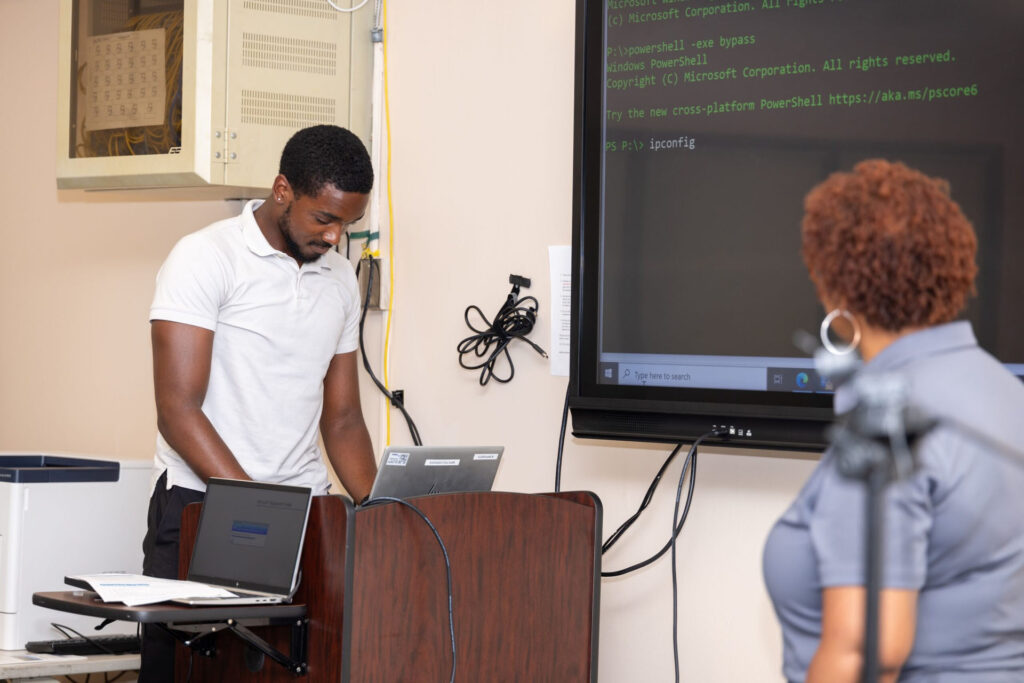 A student participates in a demonstration with a Leidos representative. The student types command line prompts into a laptop that shows the display on a large screen.