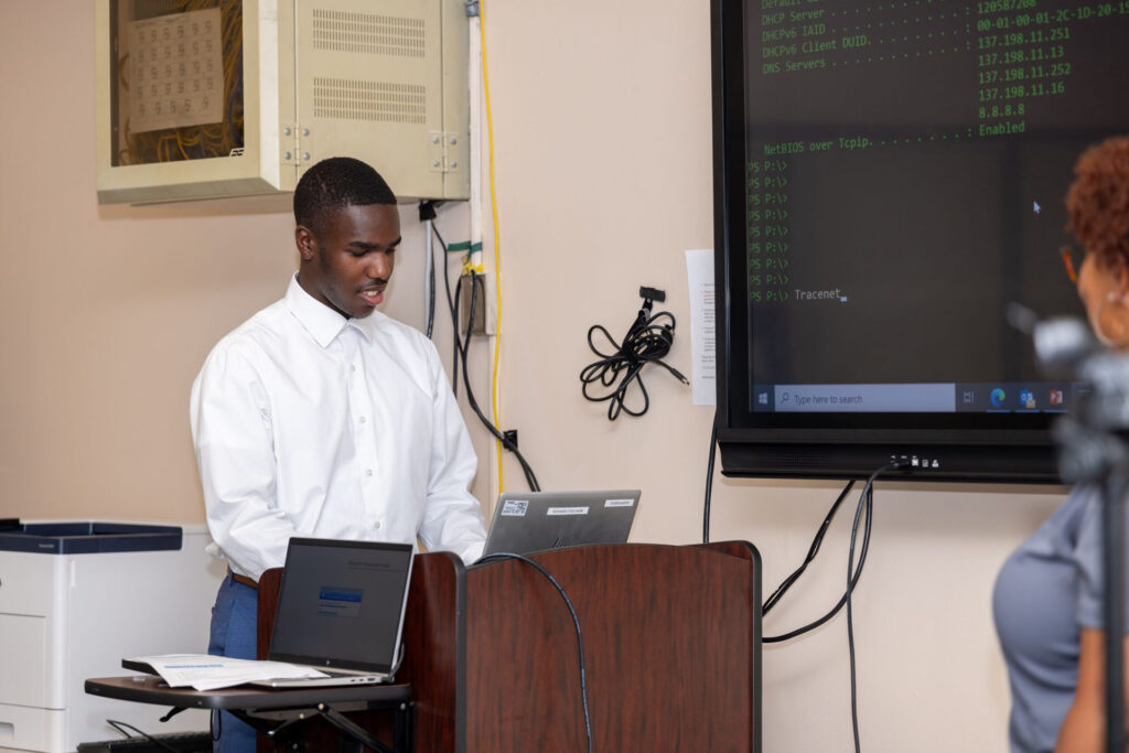 A student participates in a demonstration with a Leidos representative. The student types command line prompts into a laptop that shows the display on a large screen.