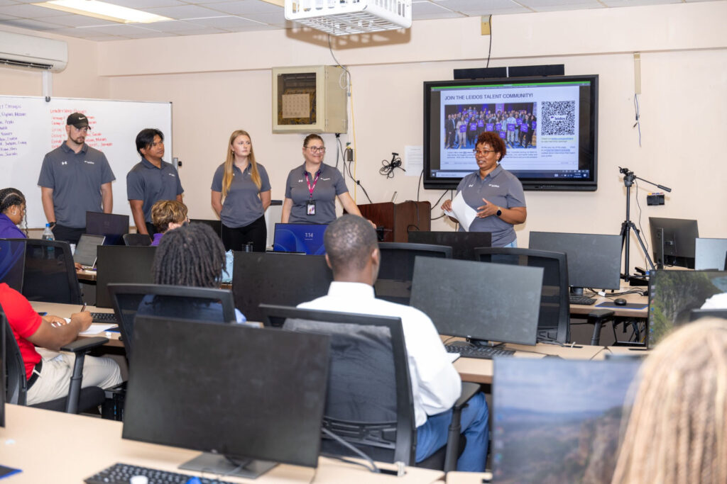 A group of Leidos representatives with a slideshow on a screen behind them. They give a presentation to students who are sitting at computers.