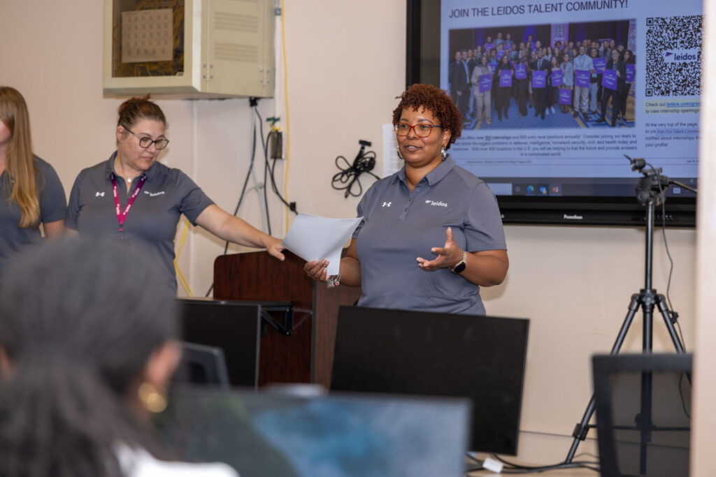 Two Leidos representatives with a slideshow on a screen behind them. They give a presentation to students who are sitting at computers.