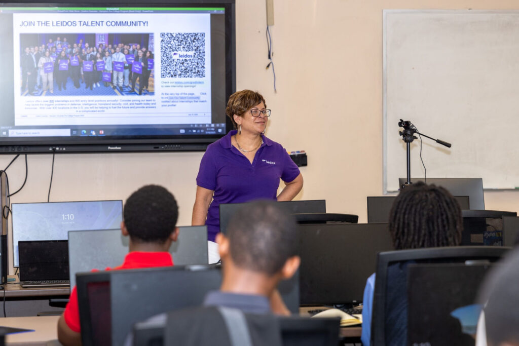 A Leidos representative with a slideshow on a screen behind her. She give a presentation to students who are sitting at computers.
