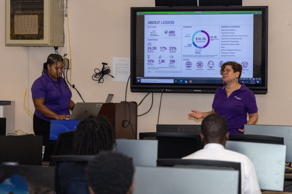 Two Leidos representatives with a slideshow on a screen behind them. They give a presentation to students who are sitting at computers.