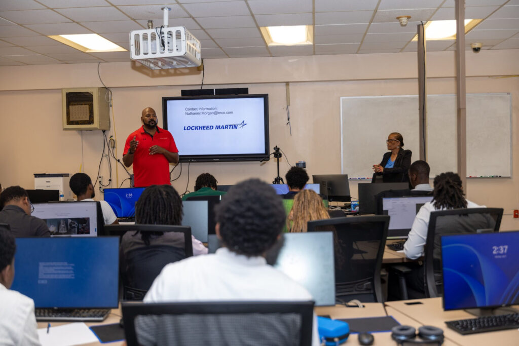 A Lockheed Martin representative gives a presentation to students with a slideshow on the screen behind him.