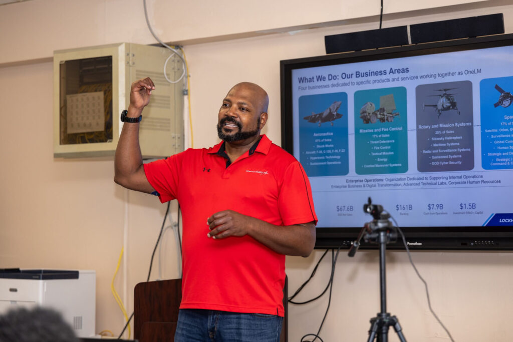 A Lockheed Martin representative gives a presentation to students with a slideshow on the screen behind him.