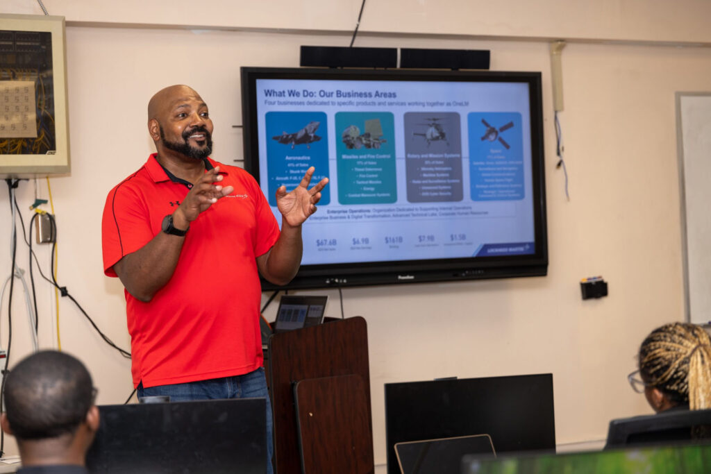 A Lockheed Martin representative gives a presentation to students with a slideshow on the screen behind him.