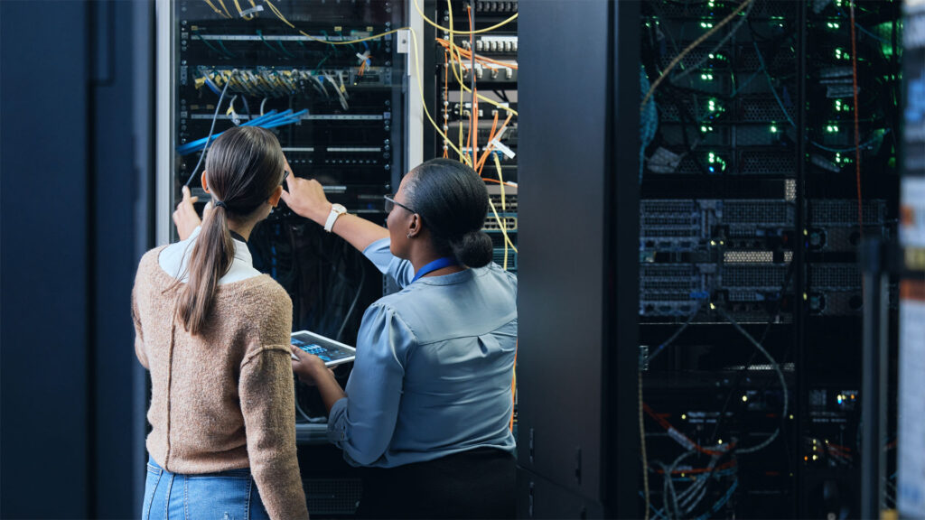 Two female students examining a network server rack with green lights.