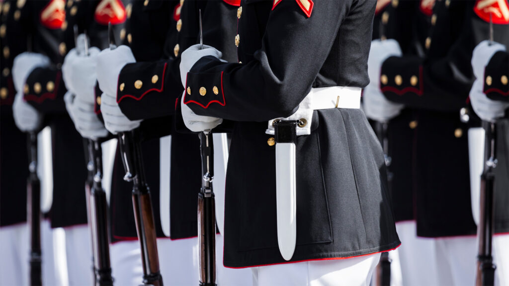 People dressed in United States Navy attire stand at attention while holding parade rifles.