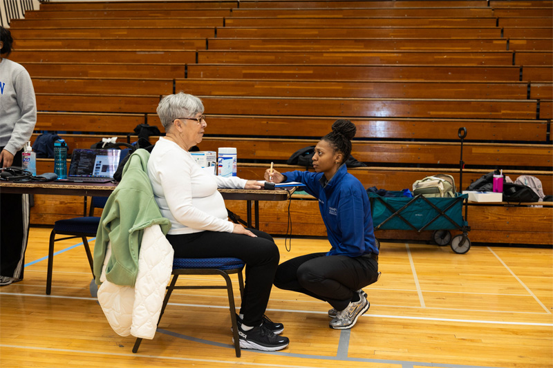 A crouched student clinician writes down a statement from a senior attendee who is sitting at a table.
