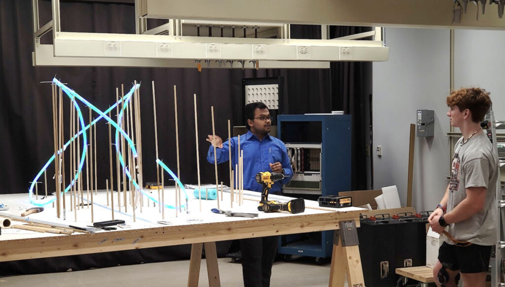 A student and professor stand around a diorama with blue lights displayed on a large, wooden table.