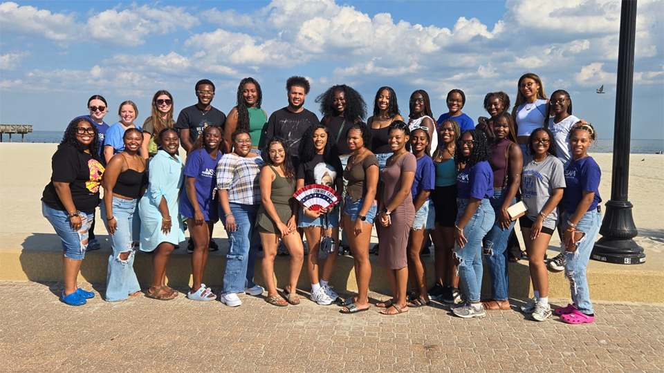 A photo of a group of students standing next to each posing for a photo on a beach.