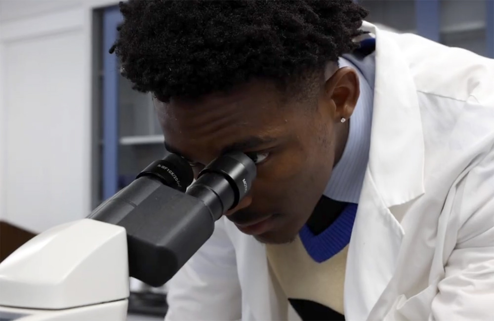 A photo of Daniel Hardy sitting in a laboratory with a microscope behind him.