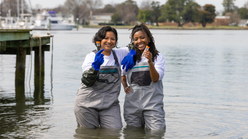 A photo of two students wearing waders standing in knee deep water while holding water samples collected in clear tubes.