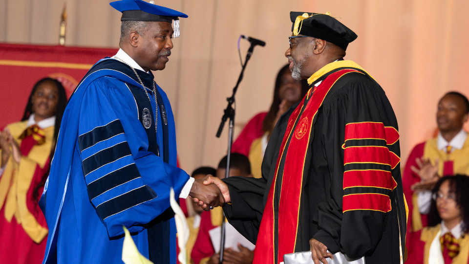A photo of Darrel K. Williams and Mark A. Brown wearing academic regalia and shaking hands.