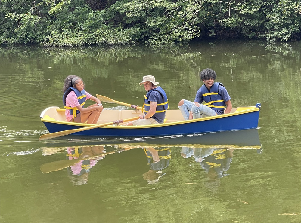 Two students on a small wooden dinghy boat with a chaperone on a lake.