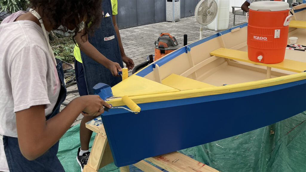A photo of a student painting the rim of a boat with yellow paint.